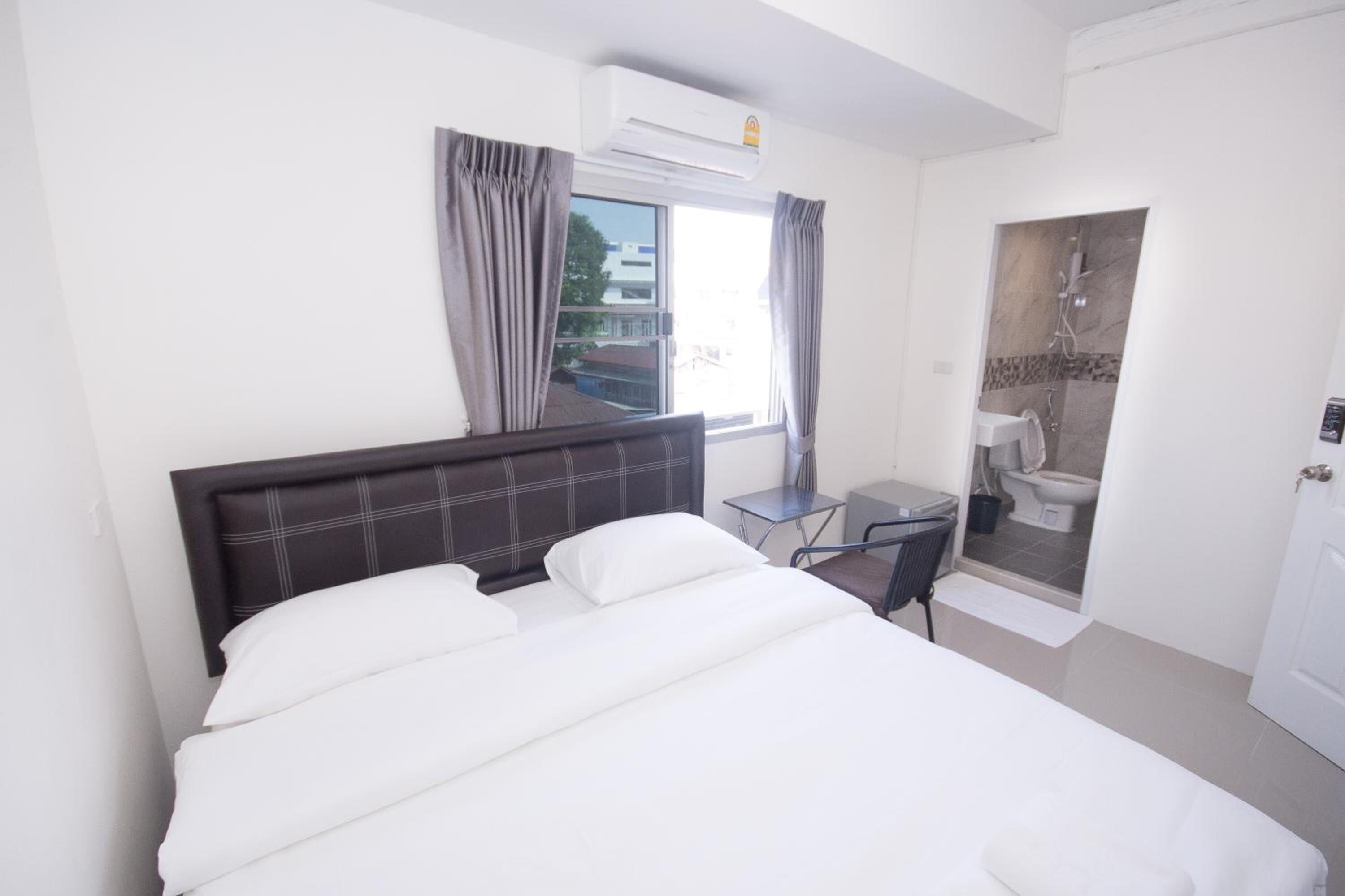 A hotel room with a bed, a desk, a chair, a window with curtains, an air conditioning unit, and a bathroom with a shower and toilet.