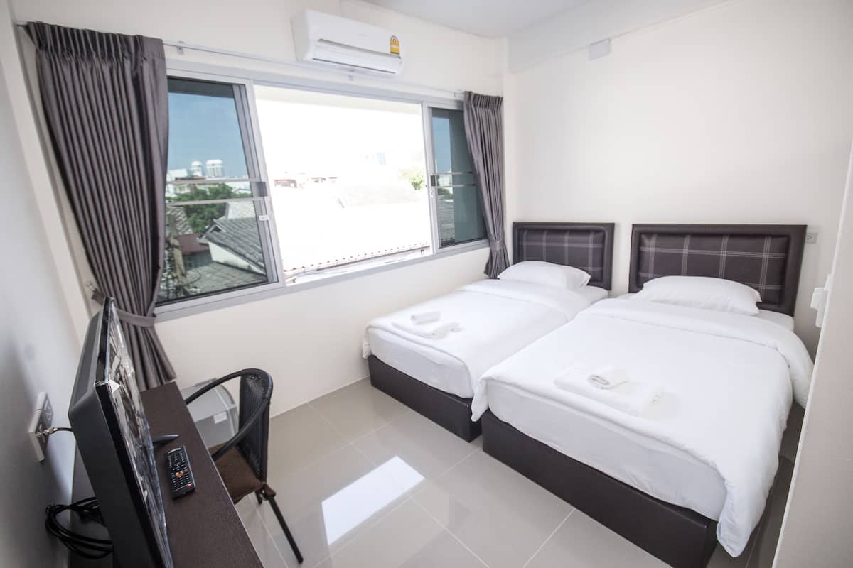 A hotel room with two beds, a desk with a chair, a window with curtains, and an air conditioning unit.