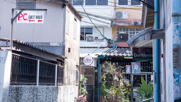 A narrow street with buildings on either side, including a guesthouse named 'Nima Guesthouse' and another with a sign 'P.C. Guest House'. There are multiple air conditioning units on the buildings, and a motorbike parked in front of the 'P.C. Guest House'. - Nina Guesthouse (Bangkok)