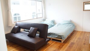 A small, bright room with a wooden floor, a bed with light blue bedding, a dark brown sofa with cushions, and a window with curtains. - Trip Spot 774 (Naha)