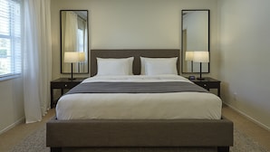 Een hotelkamer met een groot bed, twee nachtkastjes met lampen, een grote spiegel en een raam met gordijnen.