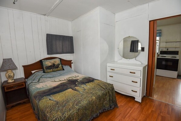 Un dormitorio con cama, mesita de noche, cómoda, espejo y una cocina visible a través de una puerta abierta.