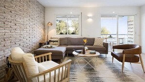 A modern living room with a brick wall, a sectional sofa, a wooden chair, a coffee table, and large windows offering a view of the outdoors. - Sitges Colonial - Downtown near the Beach (Sitges)