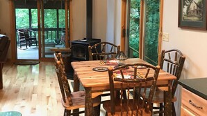 A rustic wooden dining table with four chairs, a black wood-burning stove, and a large window with curtains.