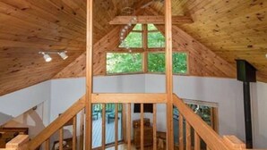 A wooden interior with a high ceiling, a central wooden staircase, and a window providing a view of greenery outside.
