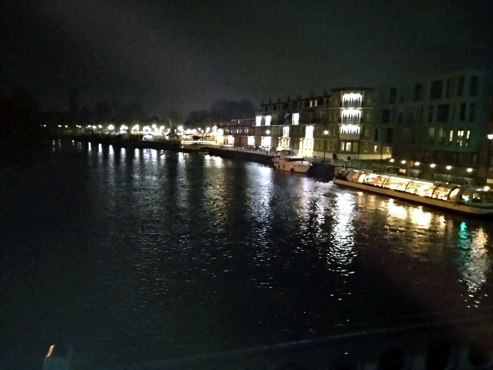 A nighttime view of a river with buildings on the right bank, illuminated by lights.