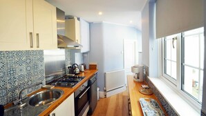 Une cuisine de taille réduite avec des meubles blancs, un évier en acier inoxydable, une cuisinière à gaz et une fenêtre équipée de stores.