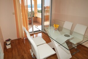 A modern dining area with a glass table and white chairs, a bowl of fruit, and a view of a balcony with a railing and potted plants. - Villa Sun (Seget)