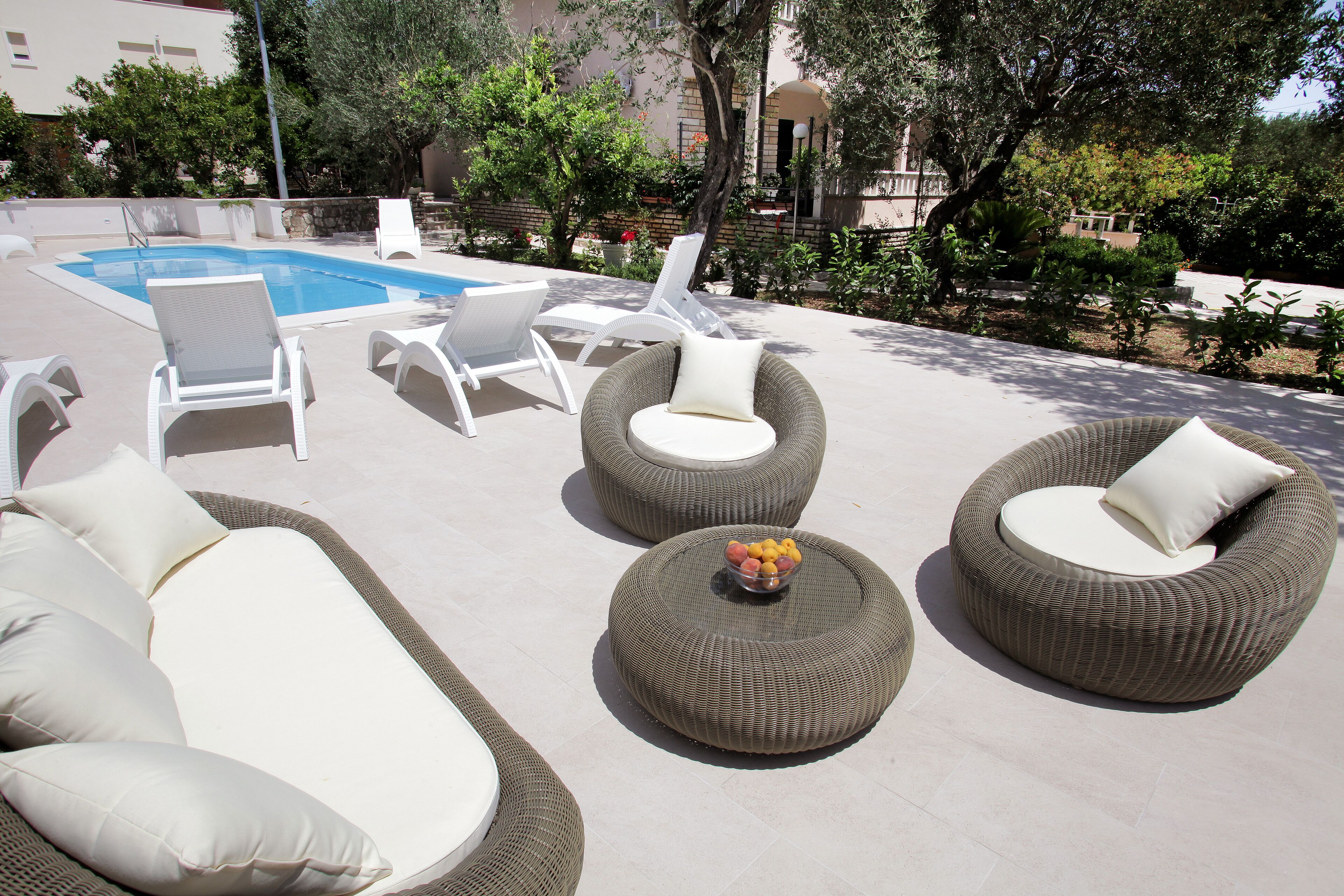 A poolside area with wicker furniture, a bowl of fruit, and a view of the pool and surrounding greenery.