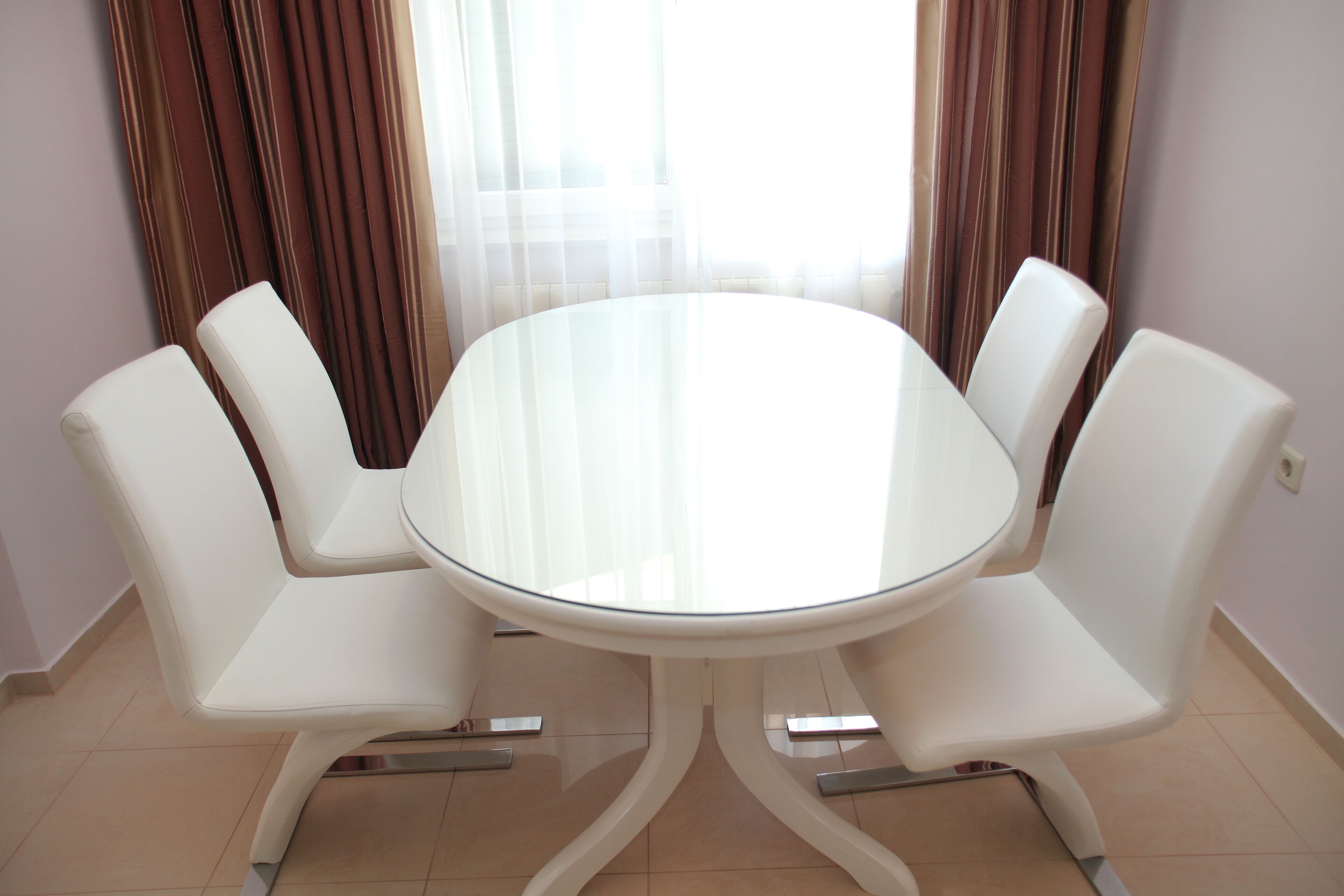 Une table ronde en verre, entourée de quatre chaises blanches, est placée dans une pièce dont le sol est recouvert de carreaux beiges et où sont accrochées des rideaux marron.