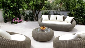 Un sofá de mimbre con cojines blancos, una mesa redonda con fruta y sillas de mimbre en un jardín.