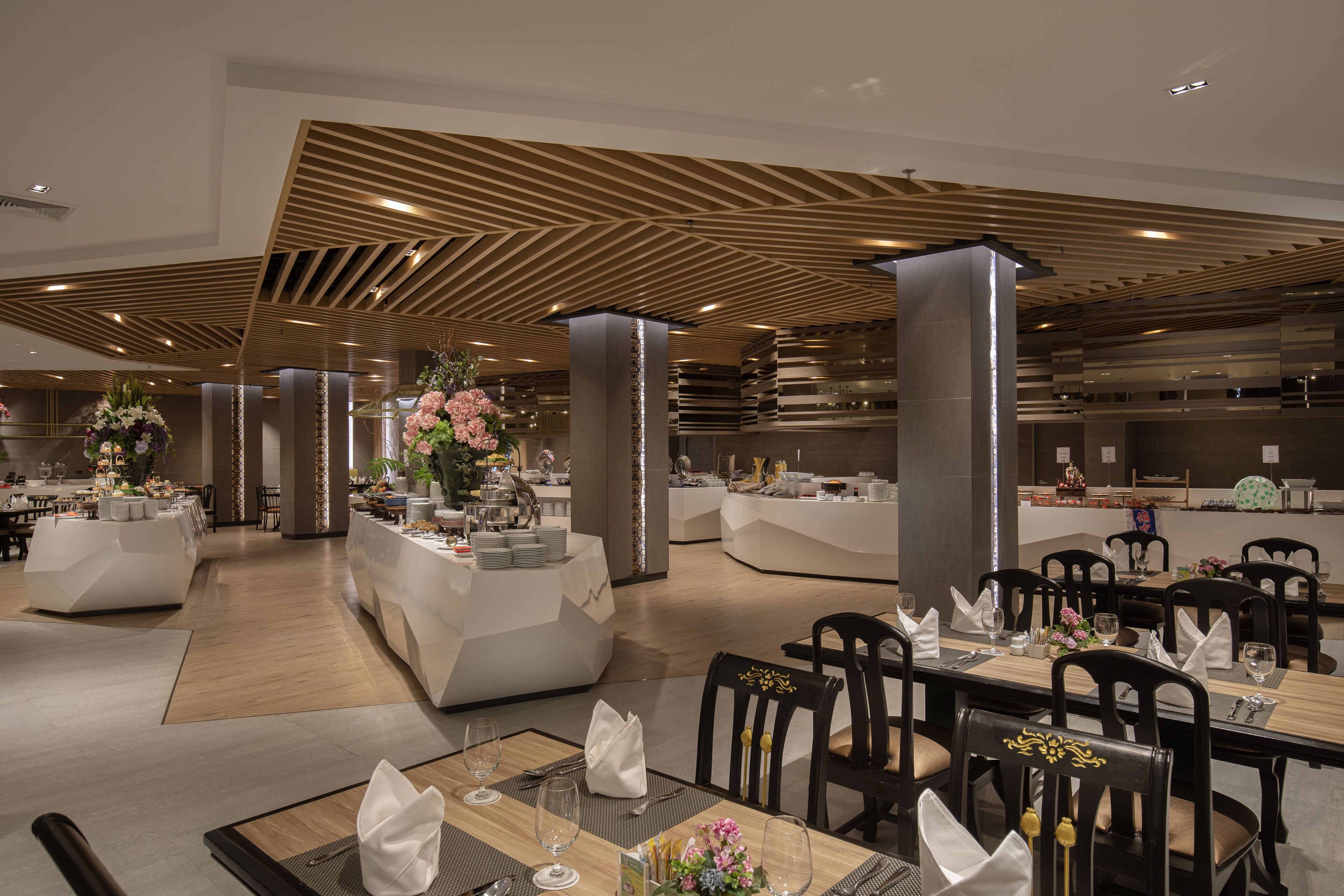 A modern dining area with geometric ceiling design, multiple dining tables set with white tablecloths, and floral centerpieces.