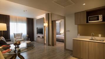 A modern hotel room with a glass dining table, a sofa, a TV, and a kitchenette.