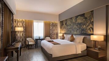 A hotel room with a large bed, two bedside tables, a desk, and a chair. There is a large tapestry on the wall and curtains with a pattern.