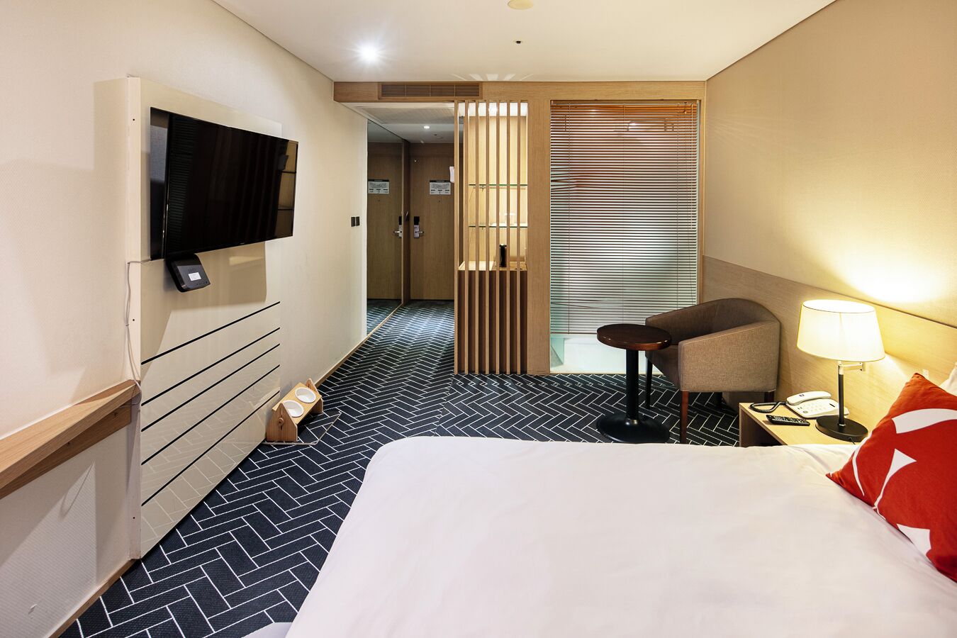 A hotel room with a bed, a TV mounted on the wall, a desk with a lamp, and a chair.