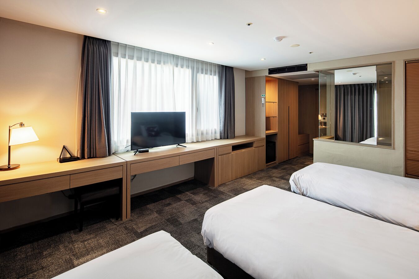 A hotel room with a bed, a desk with a TV, a lamp, and a large mirror.