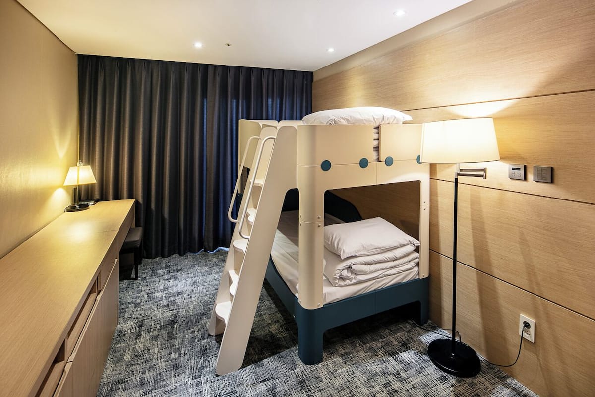 A bunk bed in a hotel room with a desk, lamp, and control panel.