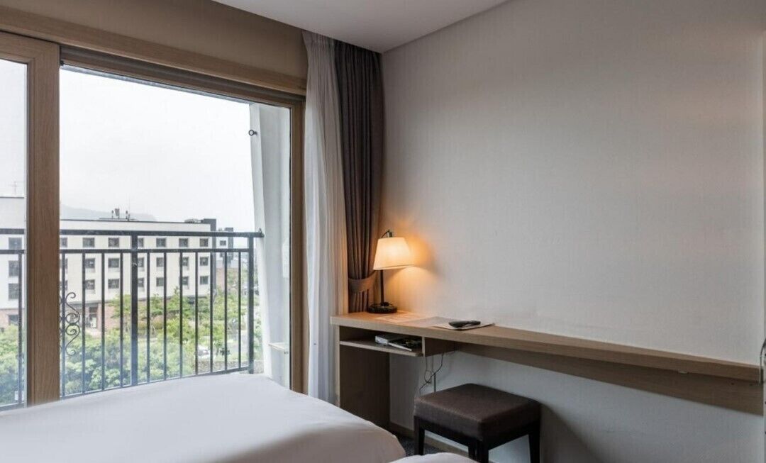 A hotel room with a bed, a desk with a lamp, a chair, and a window with a view of buildings and greenery.