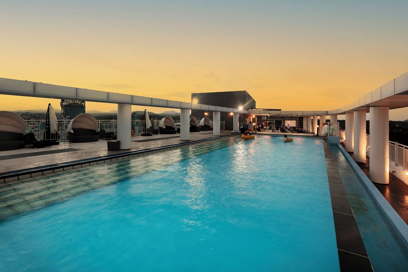 A rooftop swimming pool with clear blue water, surrounded by white pillars and umbrellas, set against a sunset sky.