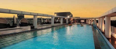 A rooftop swimming pool with clear blue water, surrounded by white pillars and umbrellas, set against a sunset sky.