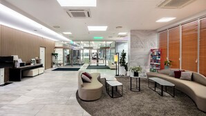 A modern lobby with a reception desk, seating area, and a large window with blinds. - The Best Jeju Seongsan Hotel (Seogwipo)
