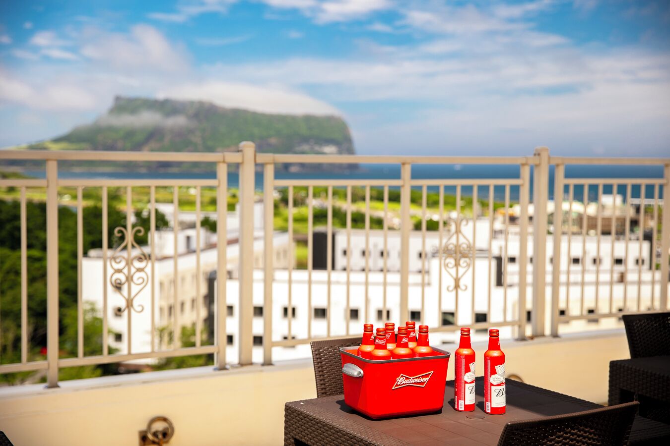 Un balcón con vista a una montaña y edificios, un enfriador rojo con botellas de Budweiser y muebles de mimbre.