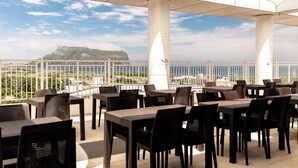An outdoor dining area with tables and chairs, a view of the sea and a distant mountain, and a white railing. - The Best Jeju Seongsan Hotel (Seogwipo)