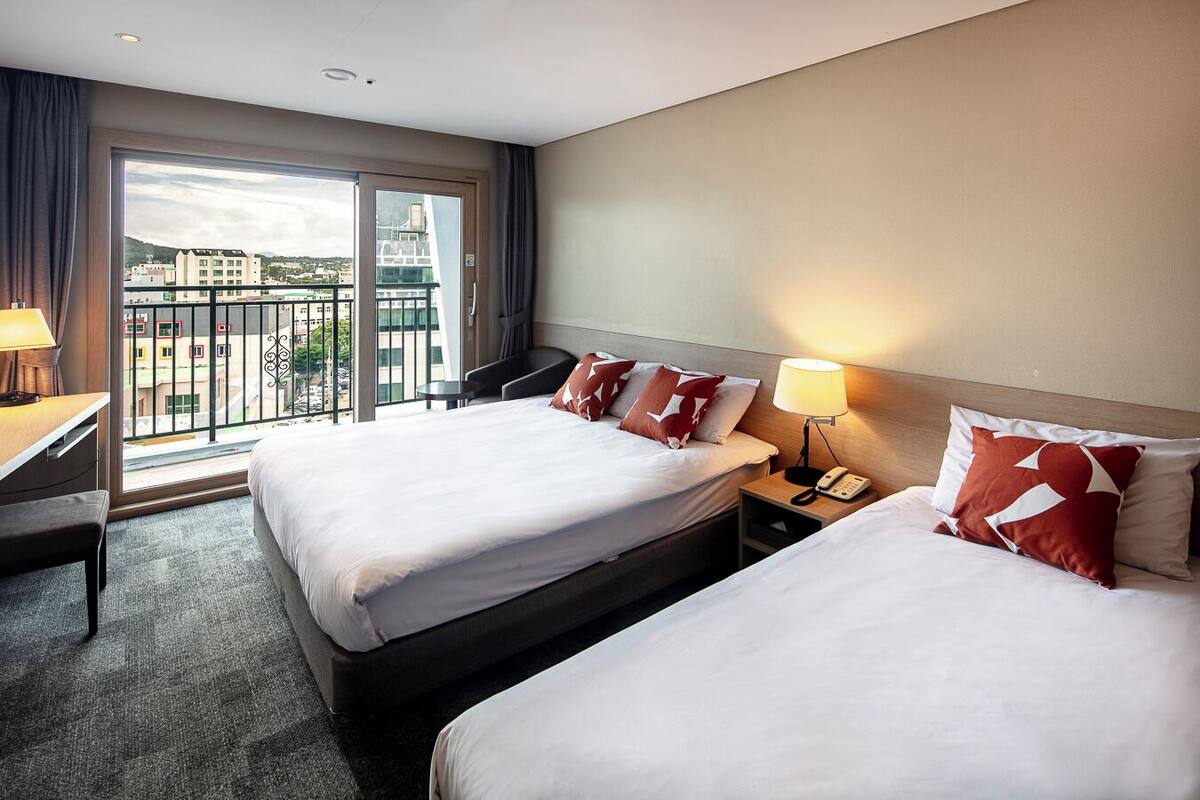 A hotel room with two beds, a desk, a chair, a phone, and a view of the city through large windows.