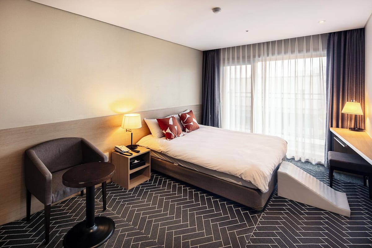 A hotel room with a bed, a desk, a chair, a nightstand, and a lamp.