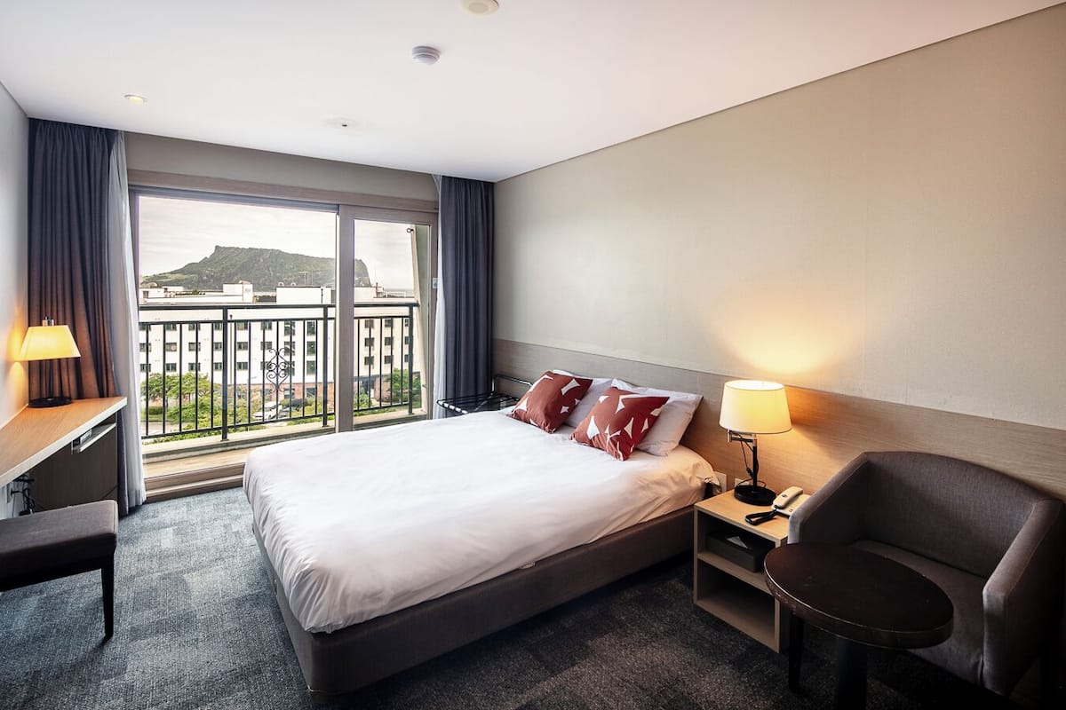 A hotel room with a large bed, a desk, a chair, and a balcony with a city view.