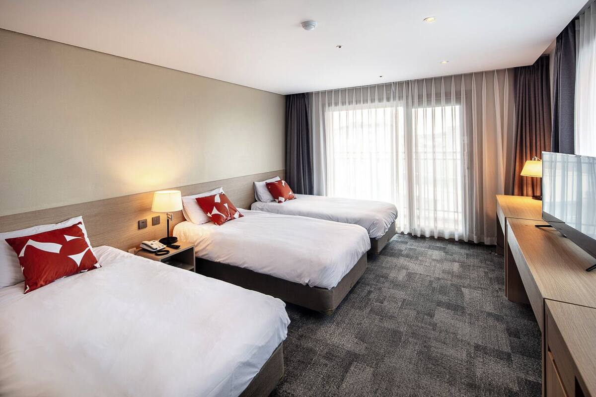A hotel room with two beds, a desk, a TV, and a large window with curtains.