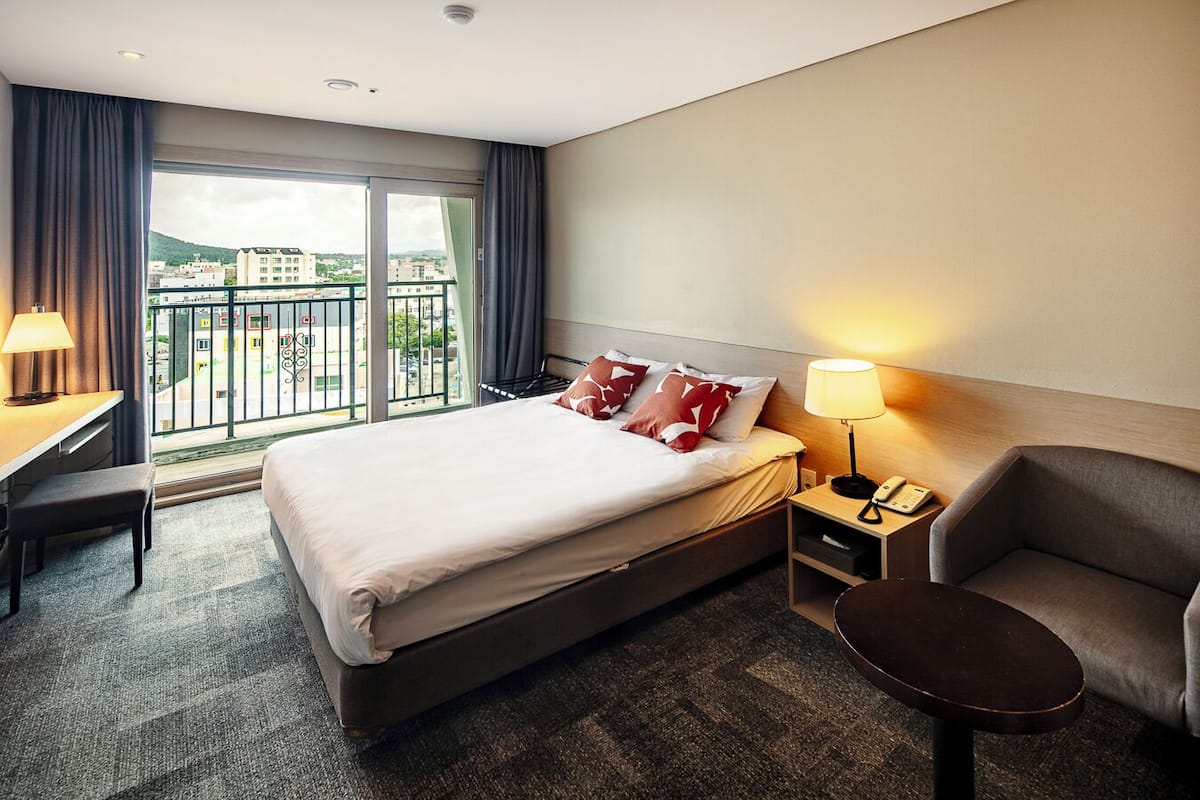 A hotel room with a bed, bedside table, chair, desk, and a view of the city through large windows.