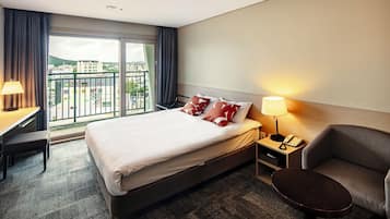 A hotel room with a bed, bedside table, chair, desk, and a view of the city through large windows.