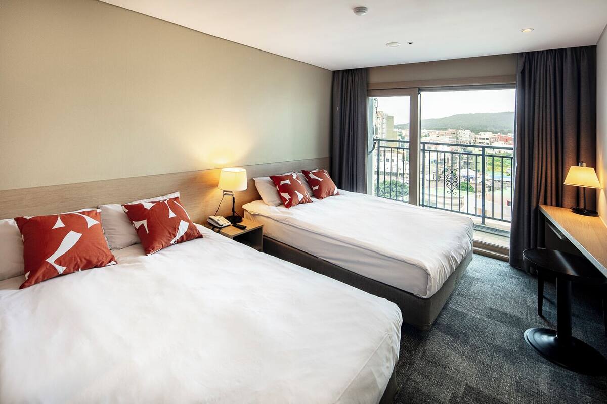 A hotel room with two beds, a desk, a chair, a phone, and a view of the city through large windows.