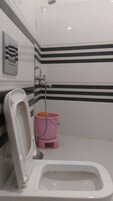 Inodoro blanco con asiento elevado, un cubo rosa y un tirador de la tapa del inodoro fijado a la pared.