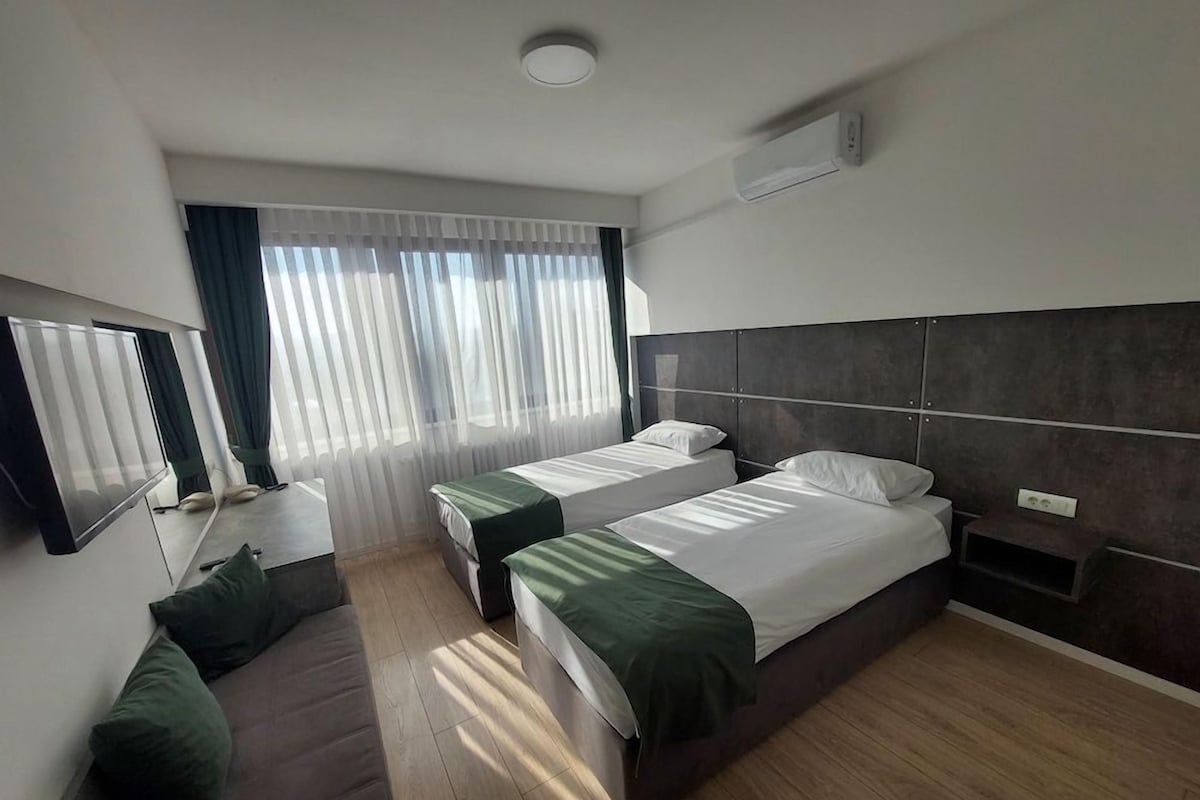A hotel room with two single beds, a sofa, a flat-screen TV, and a large window with curtains.