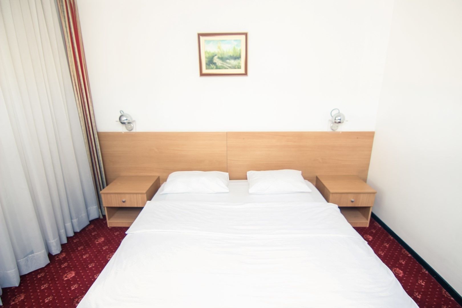 A hotel room with a large bed, two bedside tables, a nightstand, a framed picture on the wall, and a red carpet.