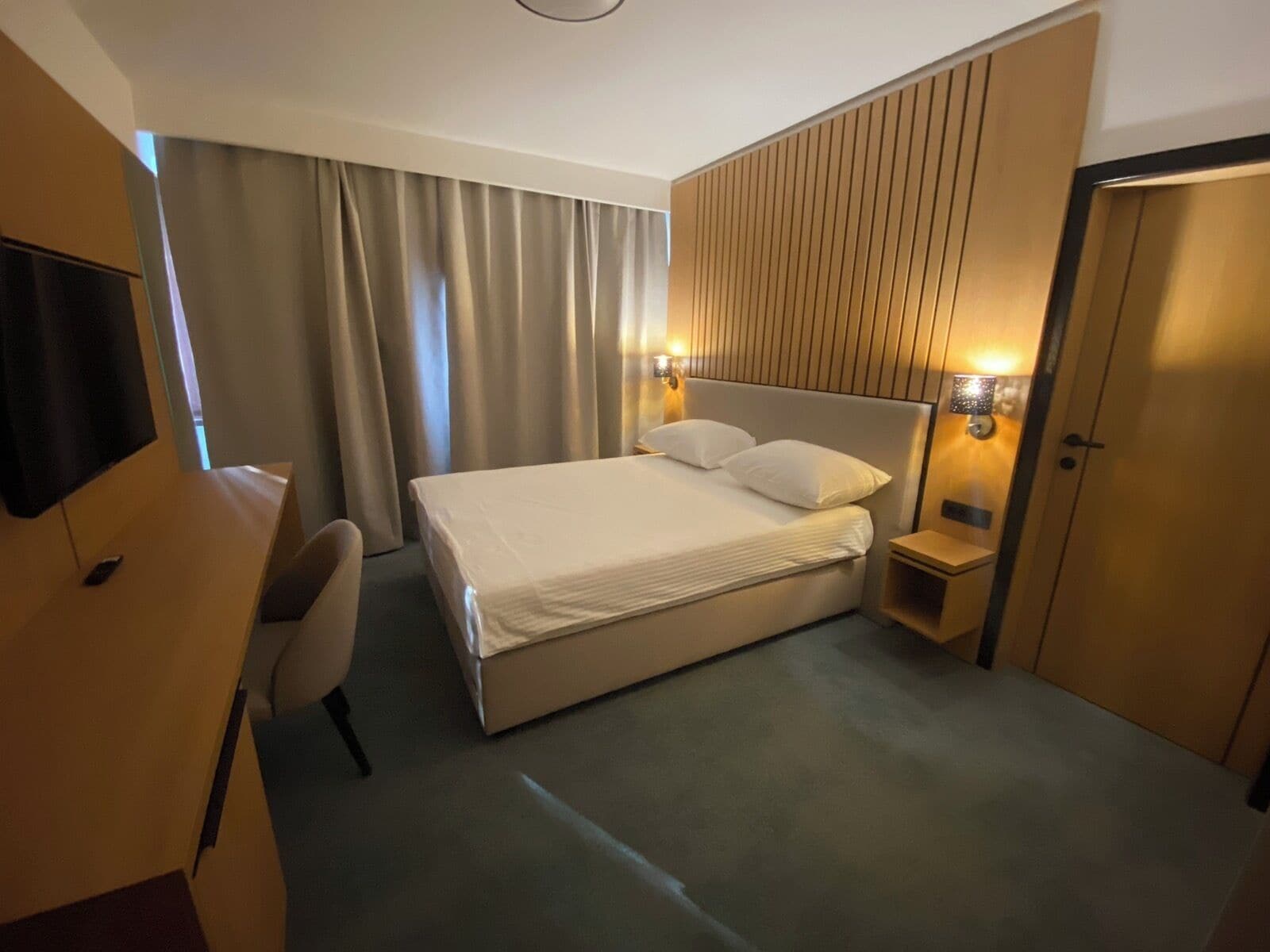 Quarto de hotel com uma cama grande, uma escrivaninha, uma cadeira, uma televisão e um guarda-roupa.