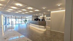 A modern reception area with a marble floor, a reception desk with a computer and a lamp, and a large glass door leading outside. - Hotel Grand (Sarajevo)