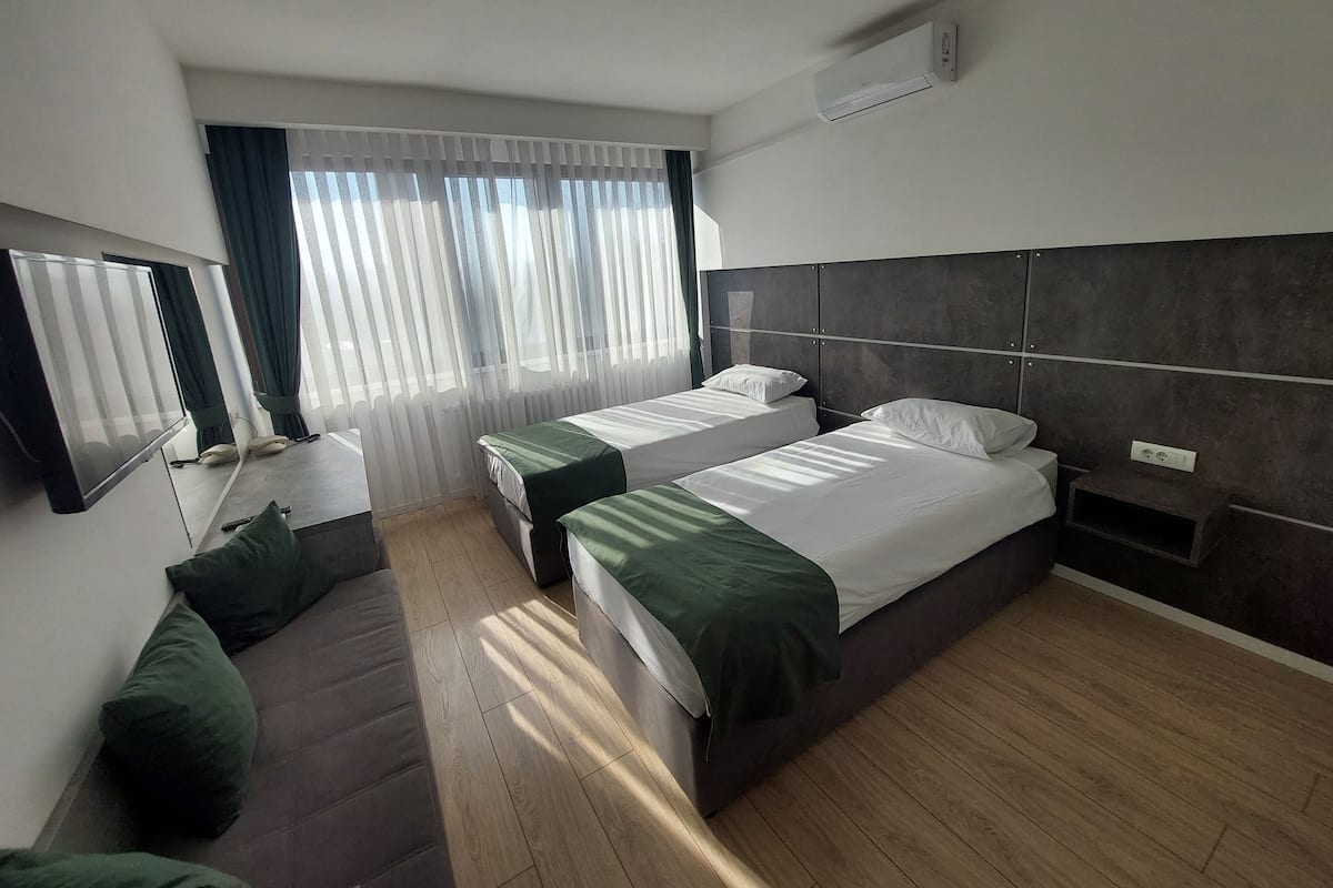 Ein modernes Hotelzimmer mit zwei Einzelbetten, einer Schlafcouch, einem Flachbildfernseher und einem großen Fenster mit durchsichtigen Vorhängen.