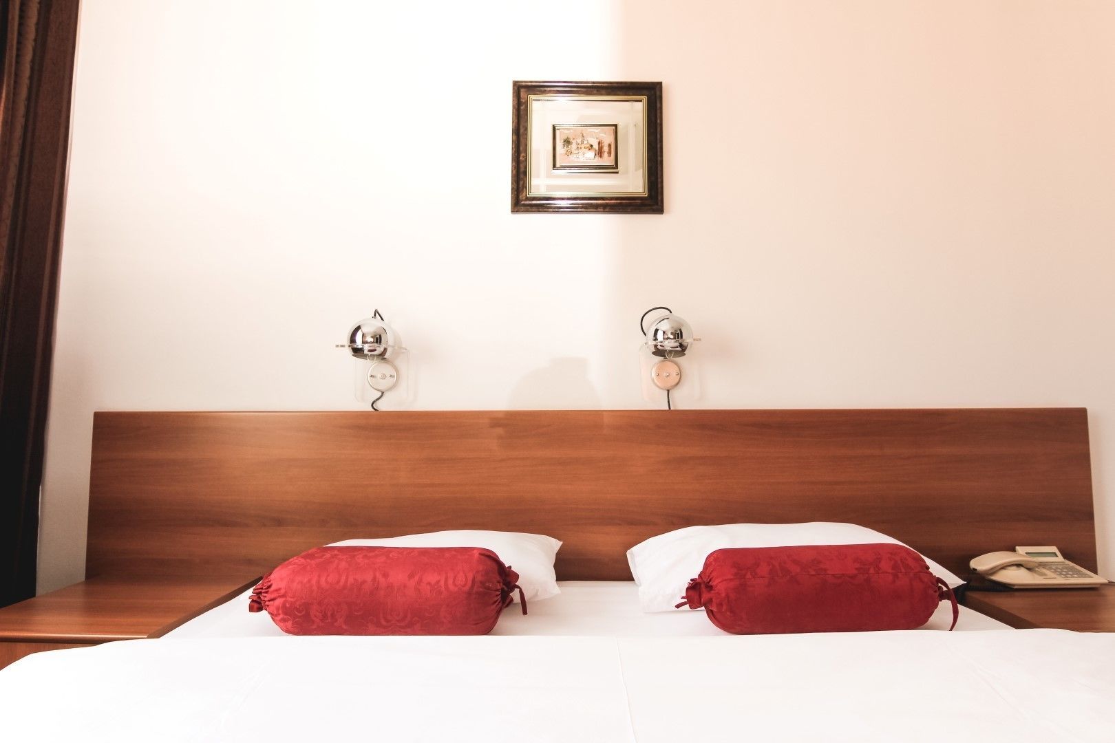 A hotel room with a wooden headboard, two red pillows, a bedside table with a phone, and a framed picture on the wall.