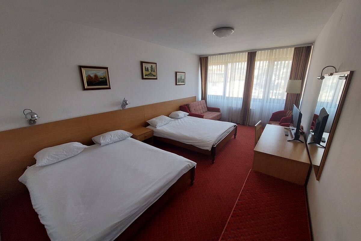 Ein Hotelzimmer mit zwei Einzelbetten, einem Schreibtisch mit Computer, einem Spiegel und einem Fenster mit Vorhängen.