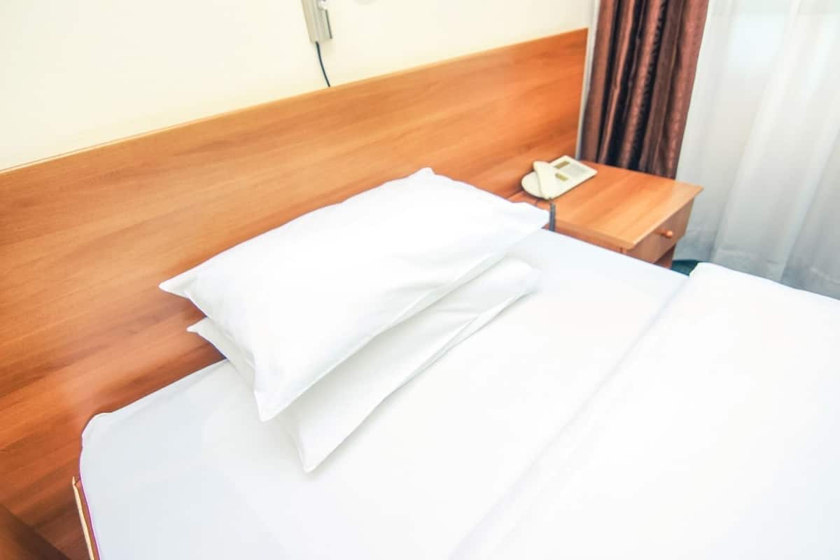 Ein ordentlich bezogenes Bett mit weißen Leinen und Kissen, ein hölzernes Kopfteil, ein Nachttisch mit Telefon und ein teilweise zurückgezogener Vorhang.