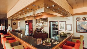 A bar area with a long counter, bar stools, and a Coca-Cola refrigerator. There are framed pictures on the wall and a wine rack in the background. - Hotel Grand (Sarajevo)