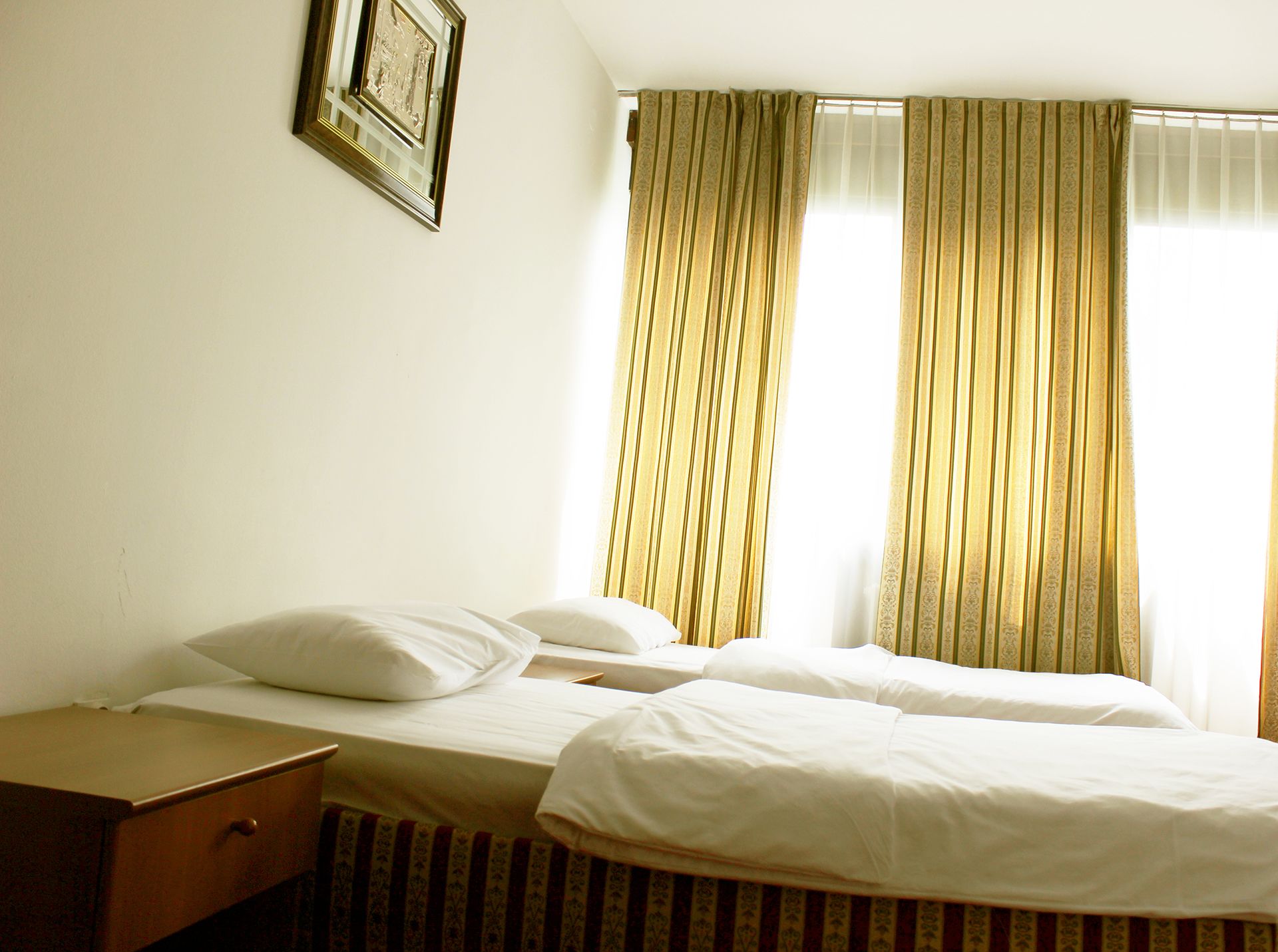 A hotel room with two single beds, a wooden nightstand, a framed picture on the wall, and curtains over the windows.