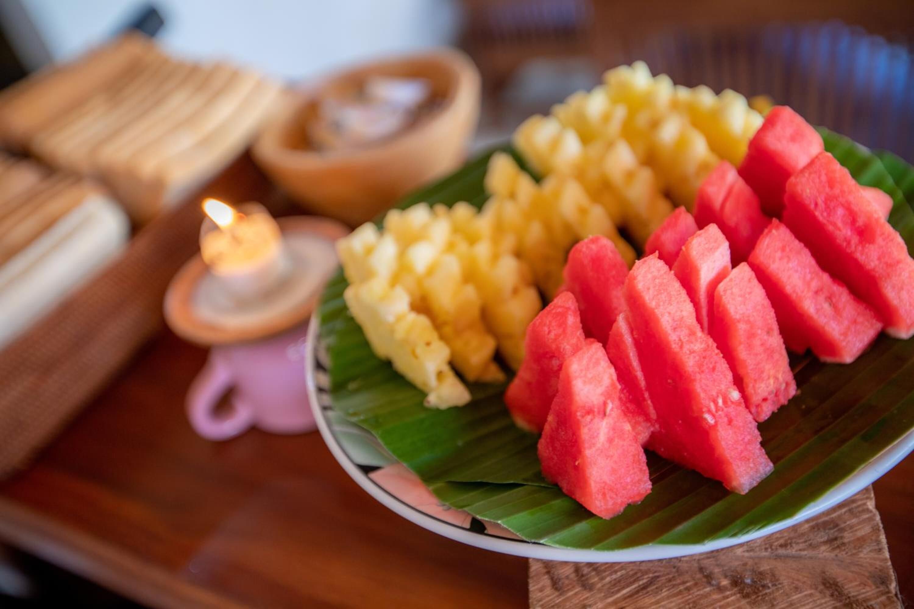 Ein Teller mit Wassermelone und Mango auf einem Bananenblatt, dazu Kerze und Holzkästen.