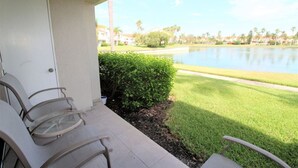 A balcony with chairs overlooking a lake, a hedge, and a row of houses in the background.
