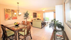 A dining area with a glass table, wicker chairs, and a bowl of fruit. There is a sofa and a plant in the background.