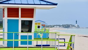 Ein Rettungsschwimmturm mit blauem Dach und grünem Unterteil, auf einem Strand mit einem Leuchtturm im Hintergrund.