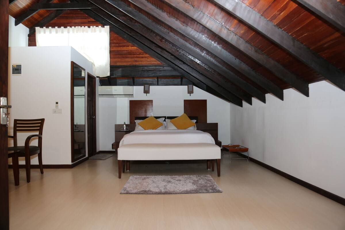 Un dormitorio con techo de madera, cama con ropa de cama blanca y dos almohadas, una silla y una pequeña mesa.